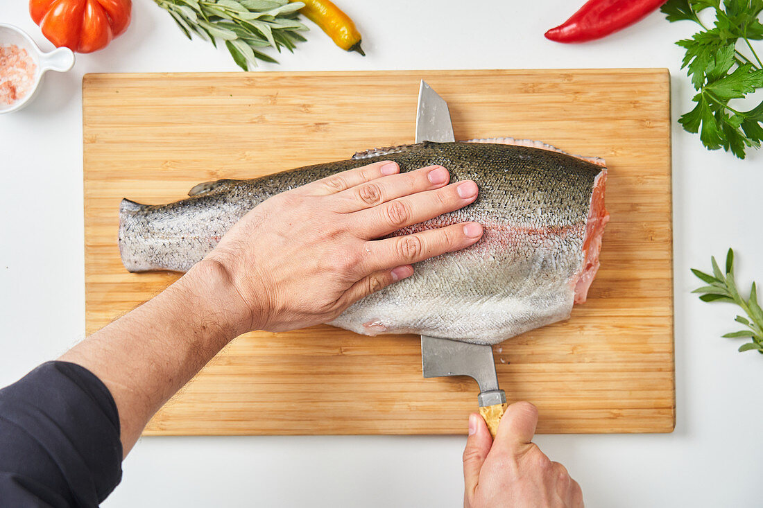 Hands cutting raw fish with knife on wooden board with fresh herbs peppers and species