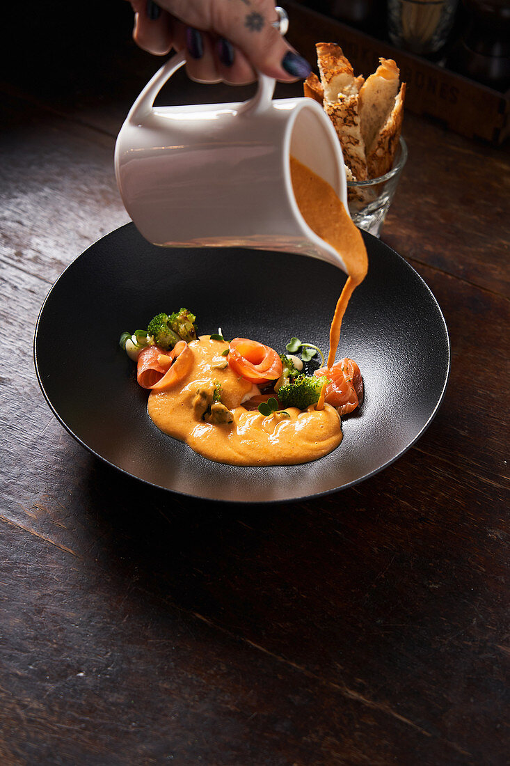 Hand of chef pouring dish with red fish and green herbs with orange sauce in stylish black bowl