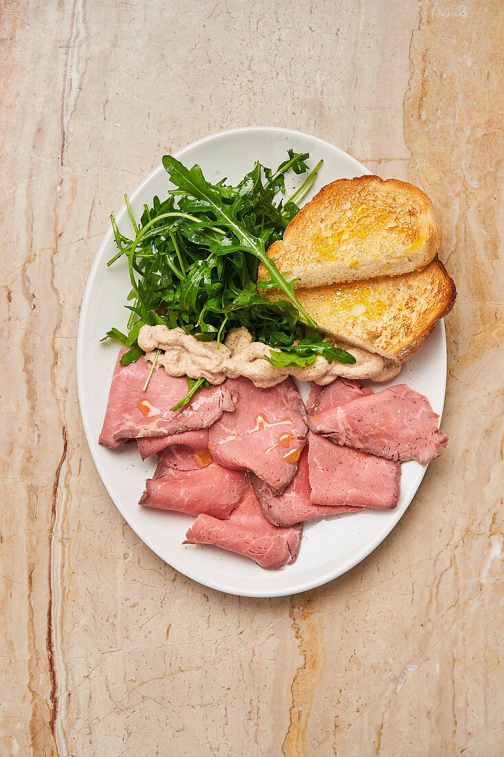 Ham with slices of roasted bread and fresh arugula in white plate on wooden table