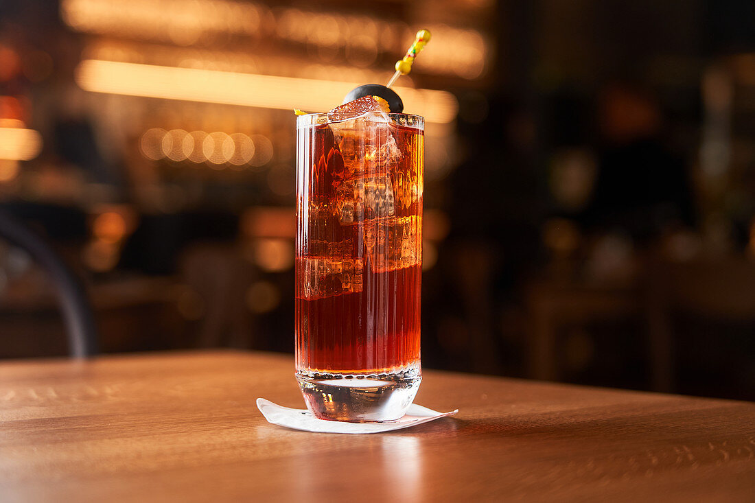 Red cocktail with ice cubes decorated with stick with black olive on wooden counter