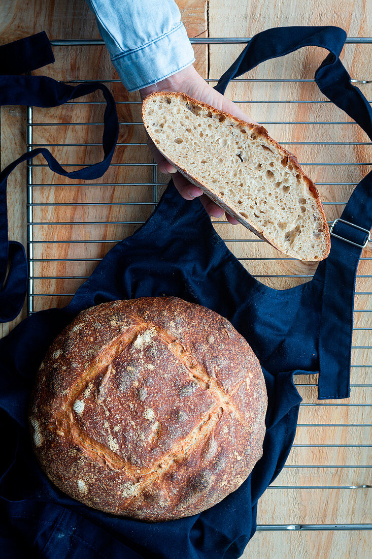 Hand holding slice of homemade sourdough bread and loaf of bread