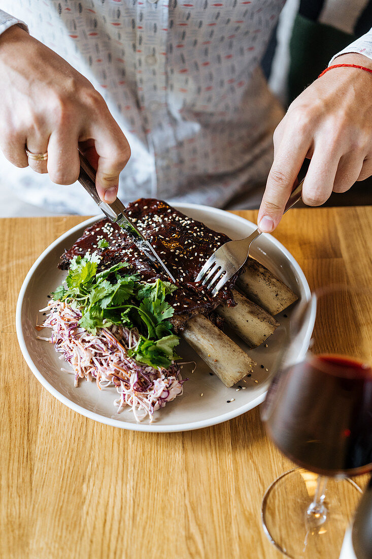 Person cutting beef ribs with a fork and knife in plate with green garnish and wine