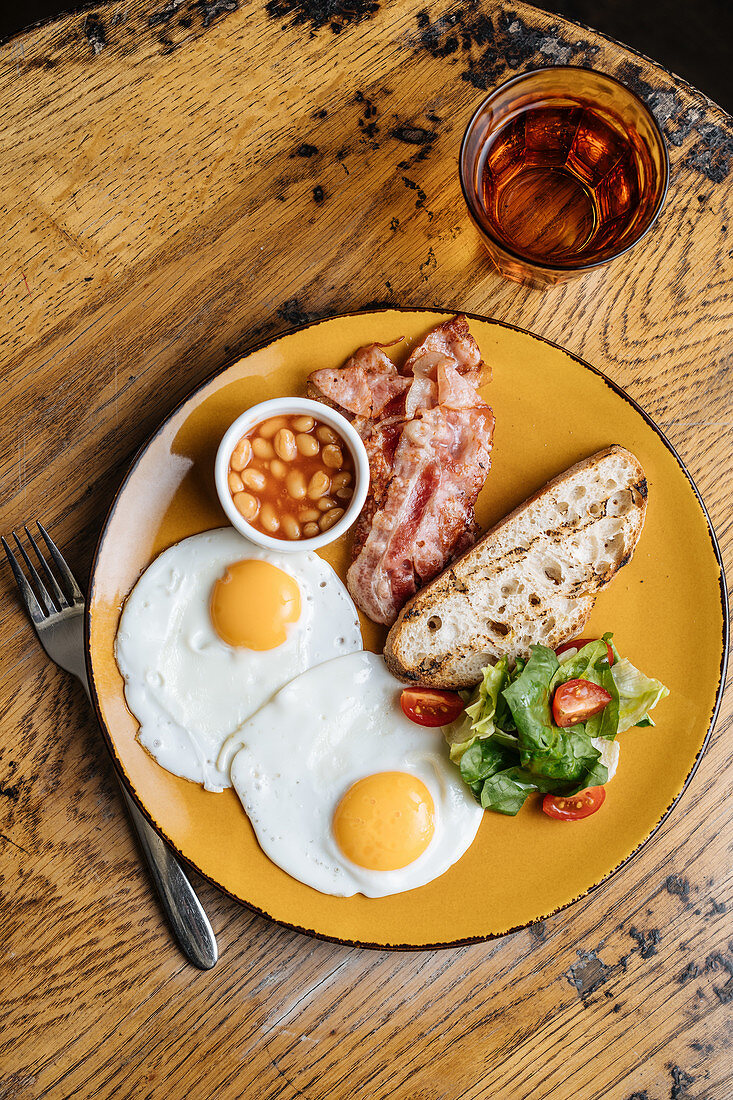 Fried eggs bacon corn in tomato greens on yellow plate in with drink