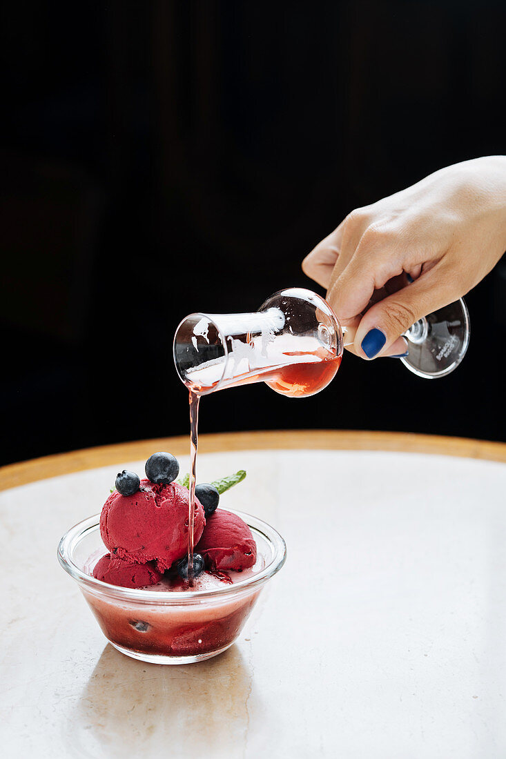 Woman pouring a red beverage on purple scoops of ice cream in bowl with fresh blueberry and mint