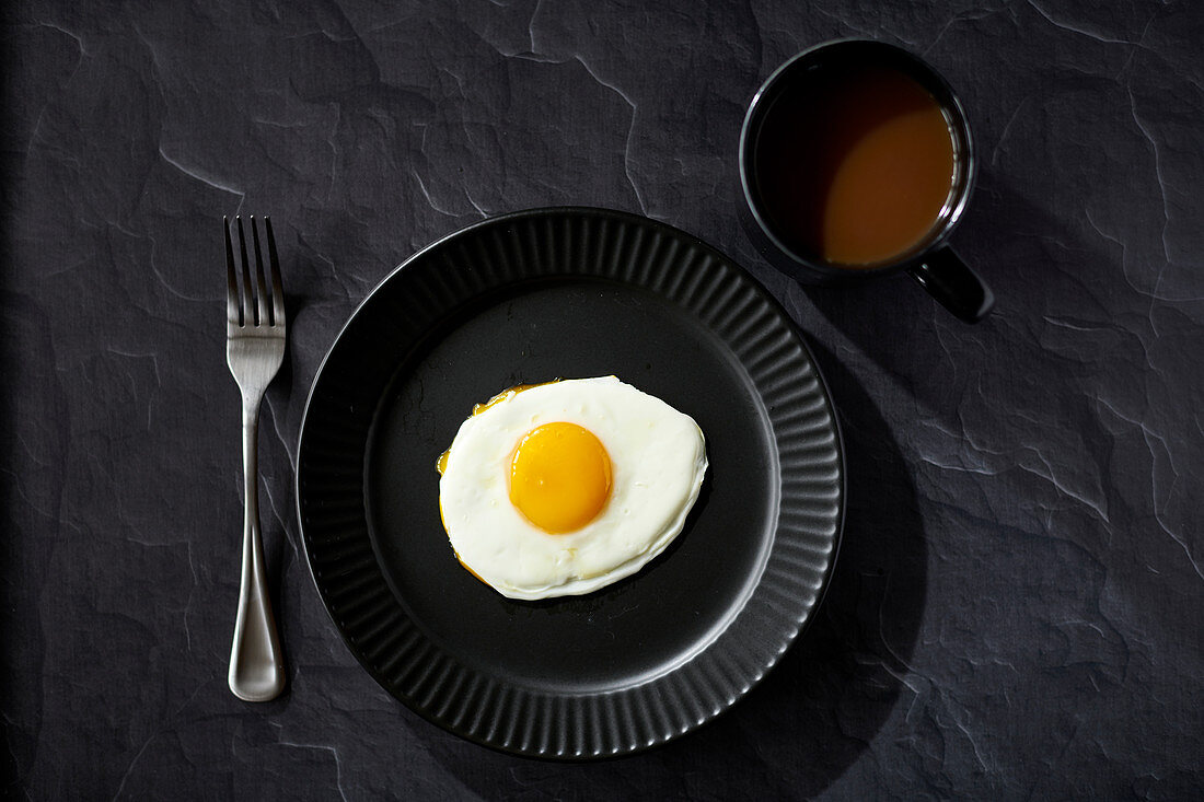 Sunnyside egg with fork and coffee on black surface with black plate and black coffee cup