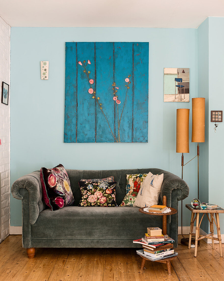 Grey velvet sofa with floral scatter cushions below blue artwork on pale blue wall