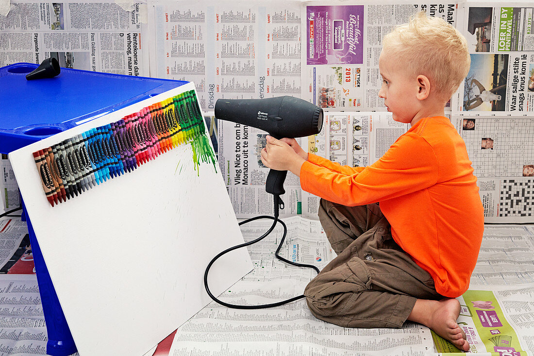 Little boy melting wax crayons on canvas using hairdryer