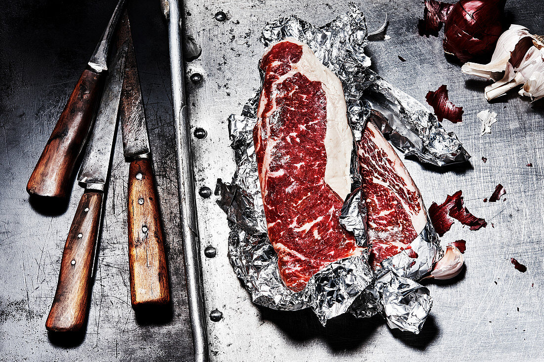 Beef steaks in aluminium foil next to old knives