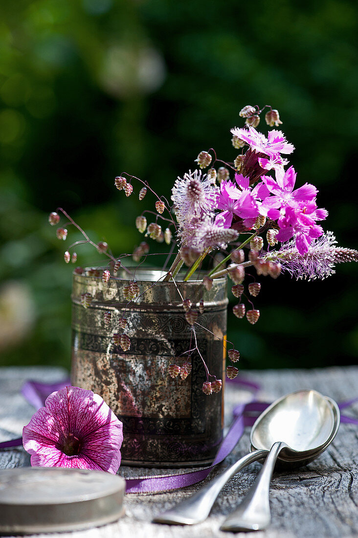 Posy of red campion, plantain flowers and wild grasses in old tea caddy next to petunia flower and spoons on table