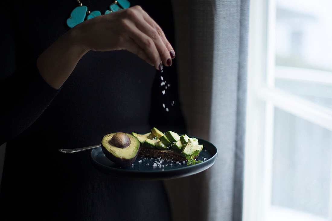 A woman standing at a window sprinkling fleur de sel onto avocado bread