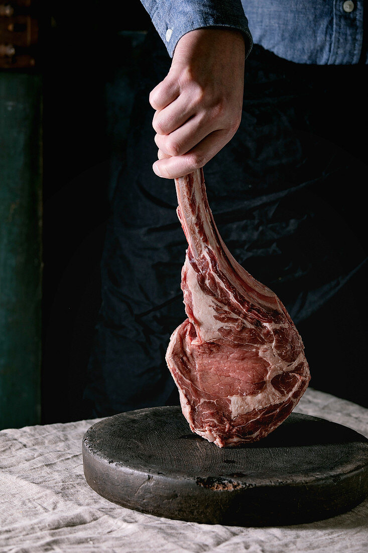 Man s hands holding raw uncooked black angus beef tomahawk steak on bones on linen table cloth. Rustic style