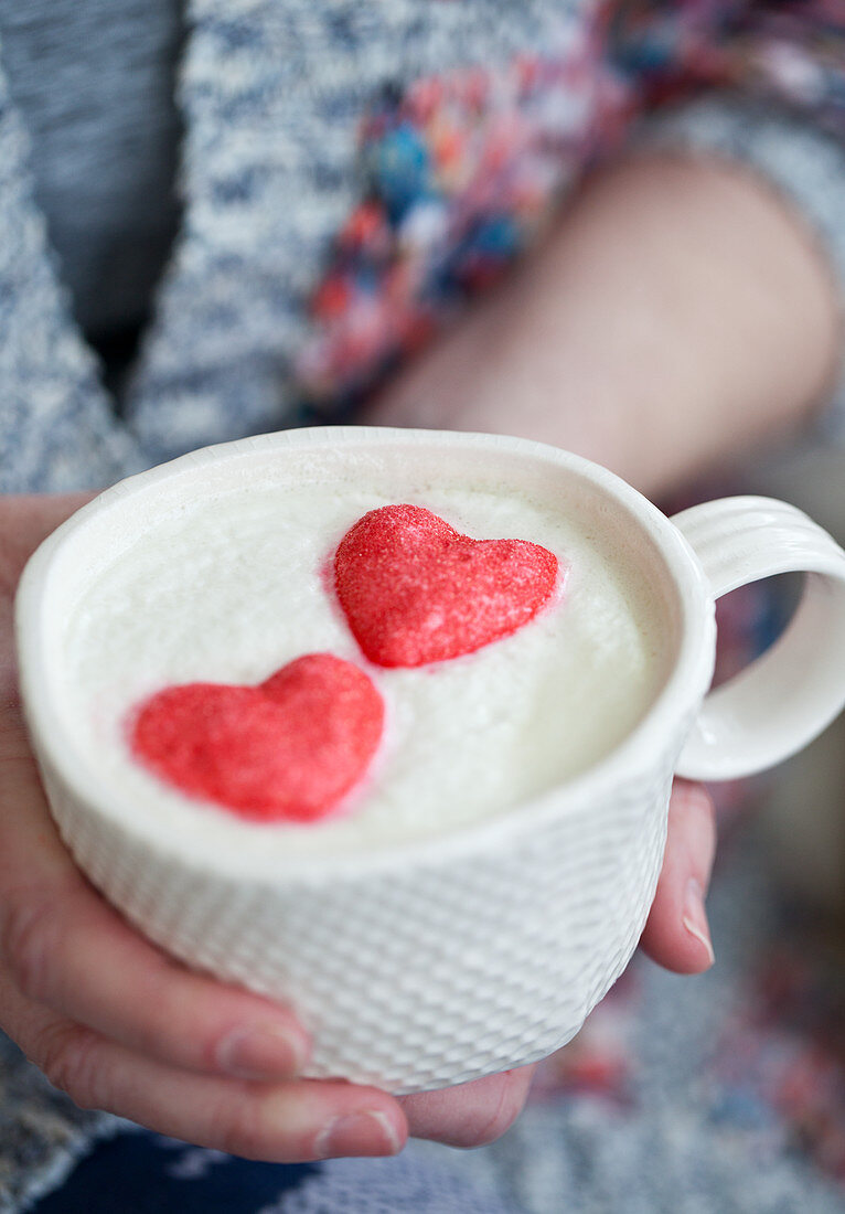 A woman wearing a mug of white hot chocolate with whipped cream and pink heart marshmallows