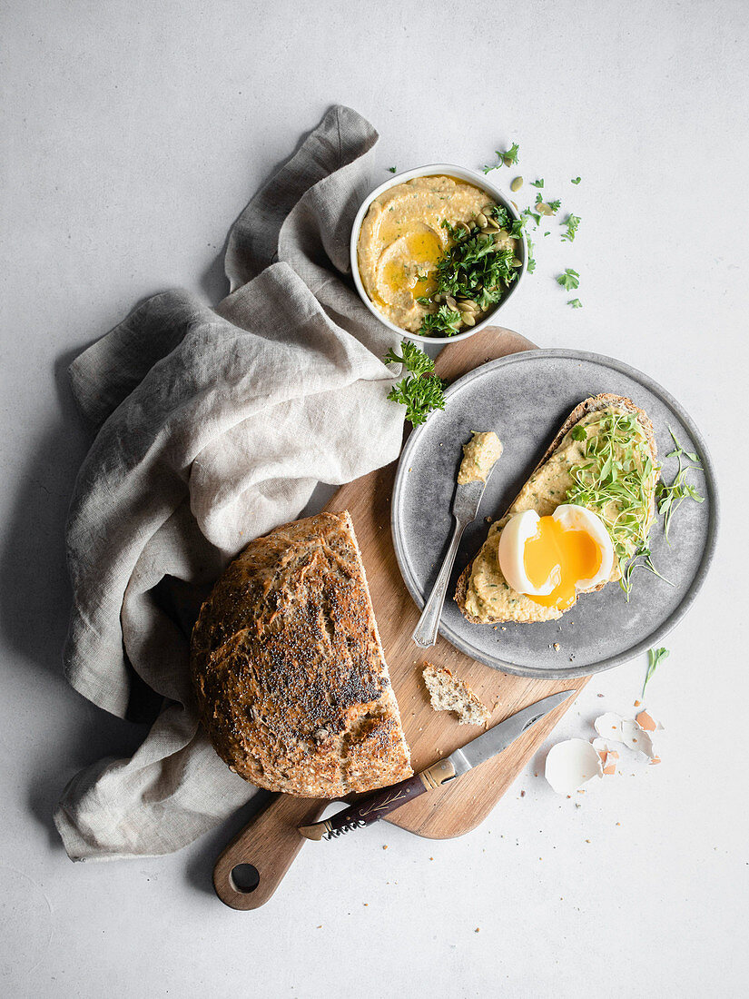 A spread of bread, hummus, and a soft boiled egg on a wood cutting board, surrounded by grey cheesecloth and garnished with parsley