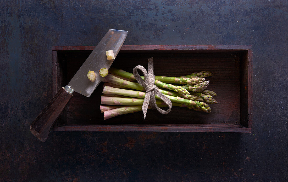 Ein Bund frischer grüner Spargel mit Messer in Holzkiste auf dunklem Untergrund