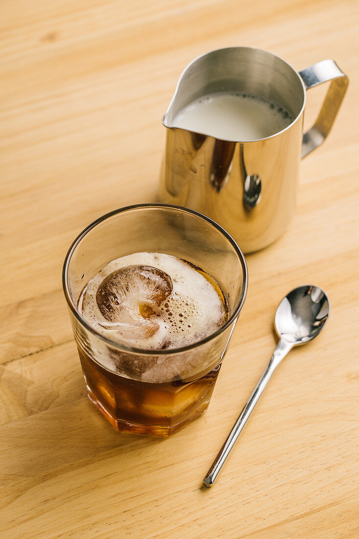 Glass with ice black coffee on wooden table with milk pitcher and spoon