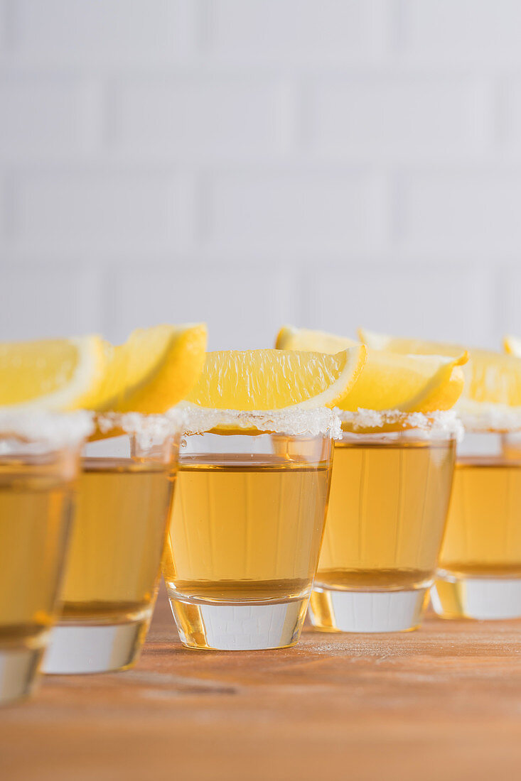 Glass sots with golden tequila and slices of lemon on wooden table