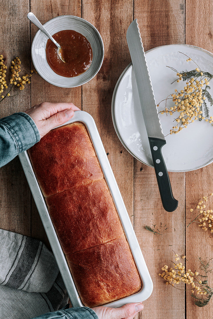 Hand with rectangle loaf of fresh Brioche bread on wooden table with bowl of jam and knife