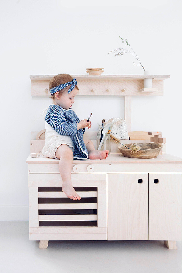 Kleines Mädchen sitzt auf Kinderküche aus hellem Holz