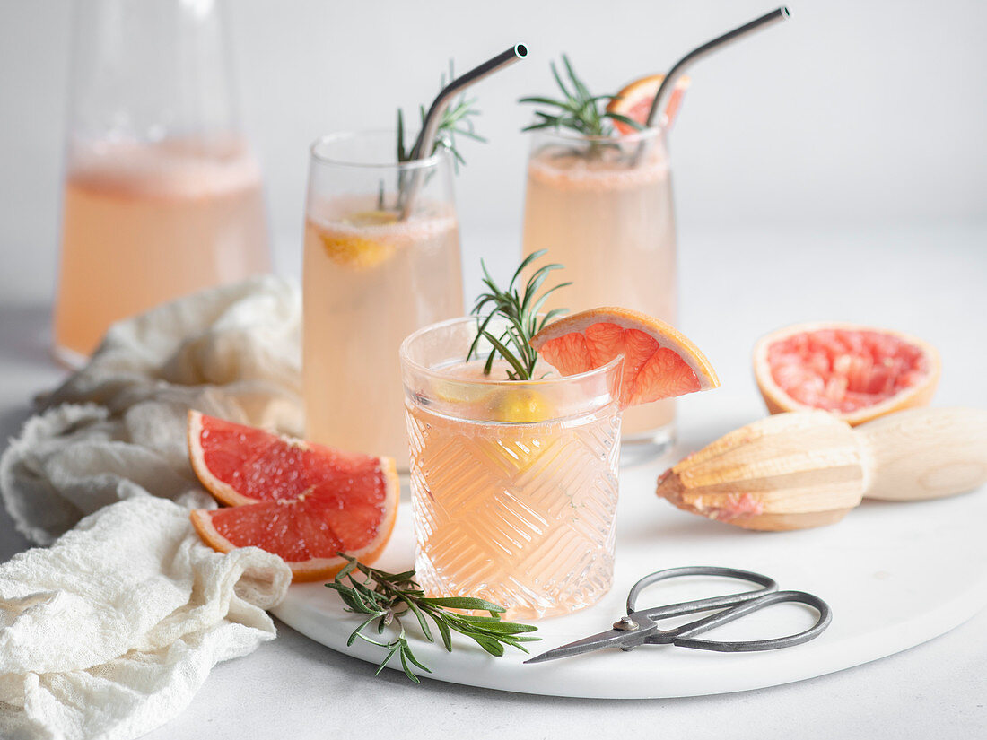 Three glasses of grapefruit cocktail on a marble tray with rosemary and grapefruit slices, pitcher and mousseline napkin