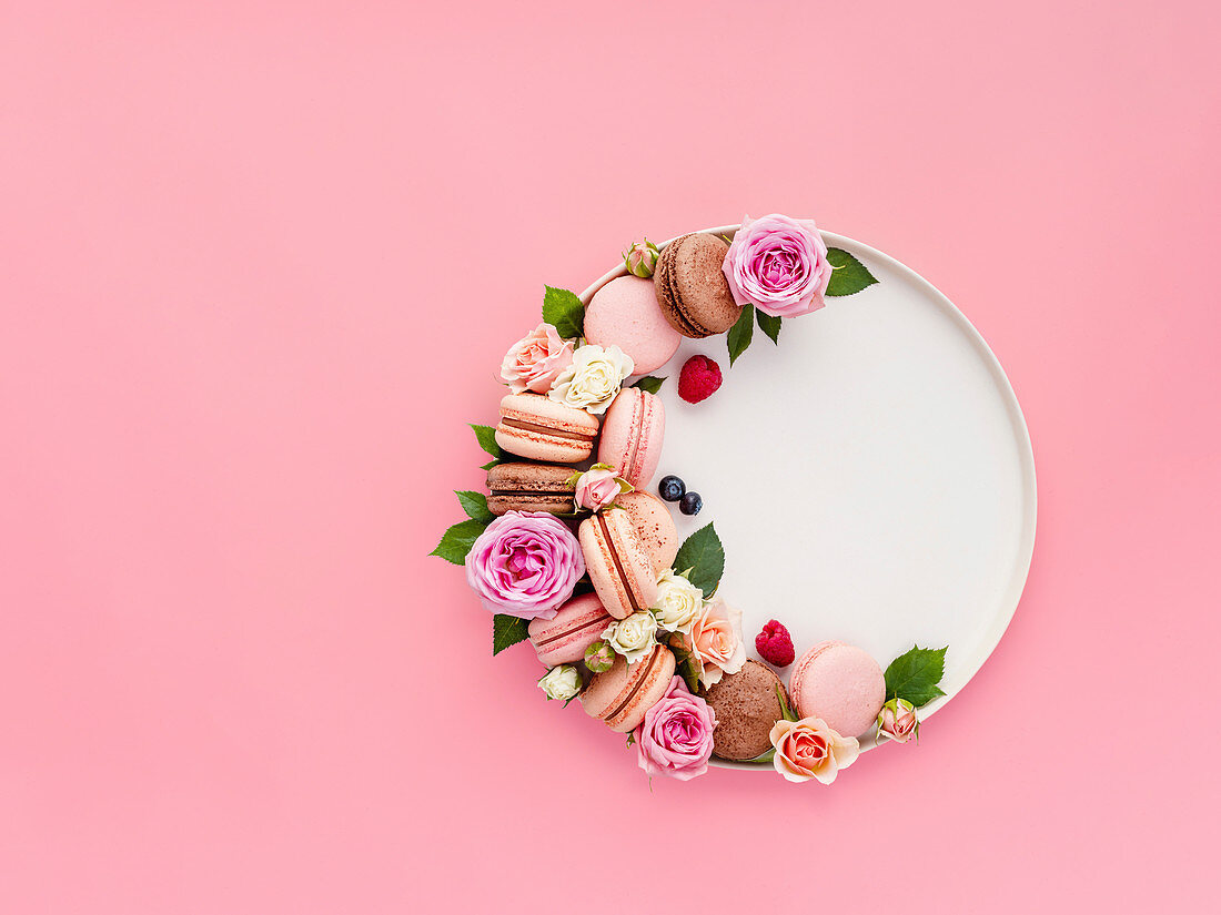Beautiful composition with french macarons, flowers and berries on craft plate over pink background