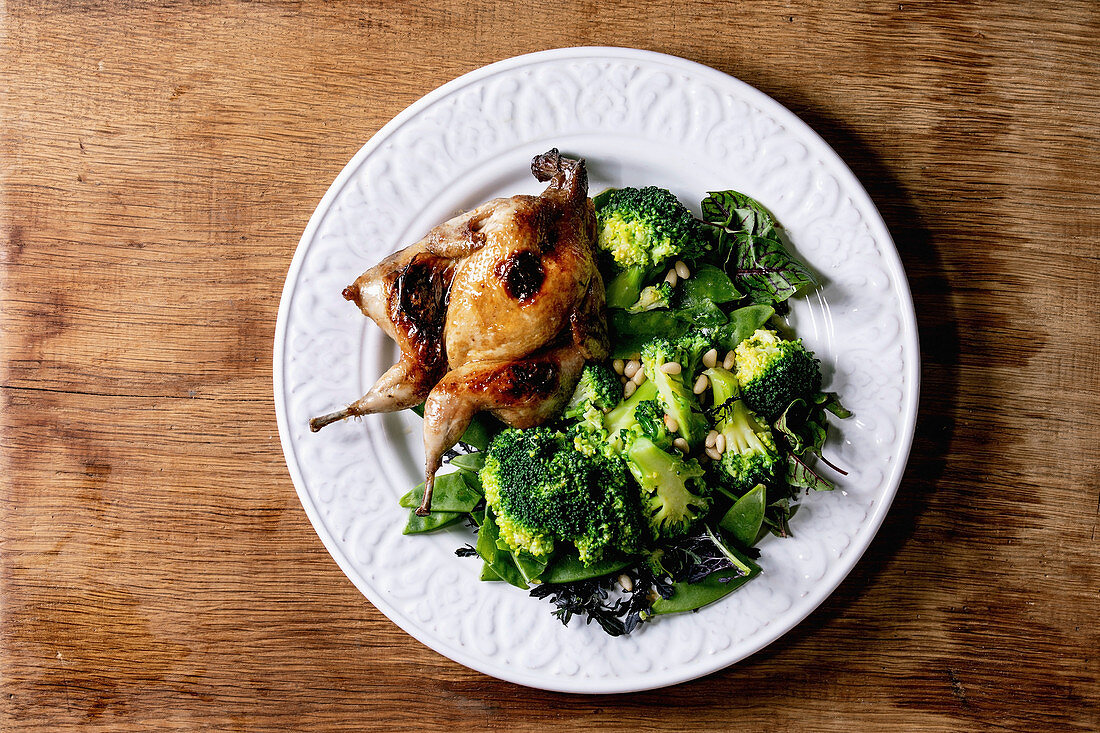 Roasted grilled butterfly quail on ceramic plate with garnish green salad and broccoli, pepper mill and olive oil