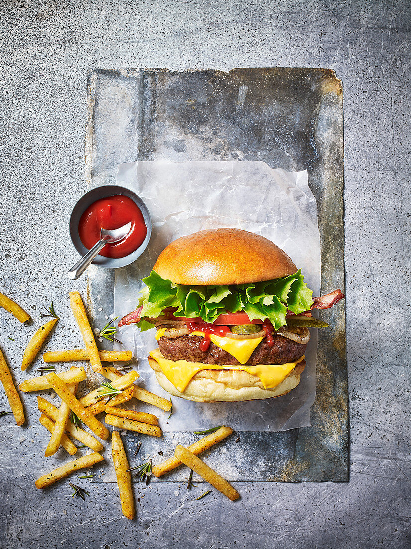 A beef burger with bacon, fries and tomato sauce against a light background