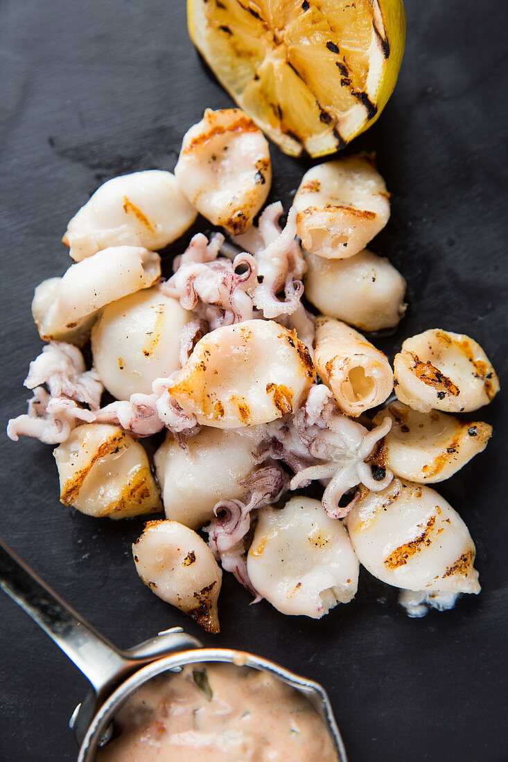 Heap of delicious grilled squids and small octopuses placed near lemon and sauce on table