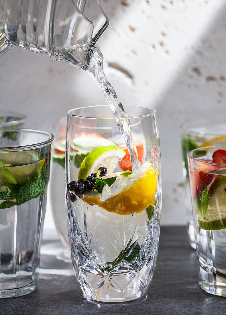 Various glasses of water filled with fruit, mint, and ginger with water being poured from a pitcher into one glass