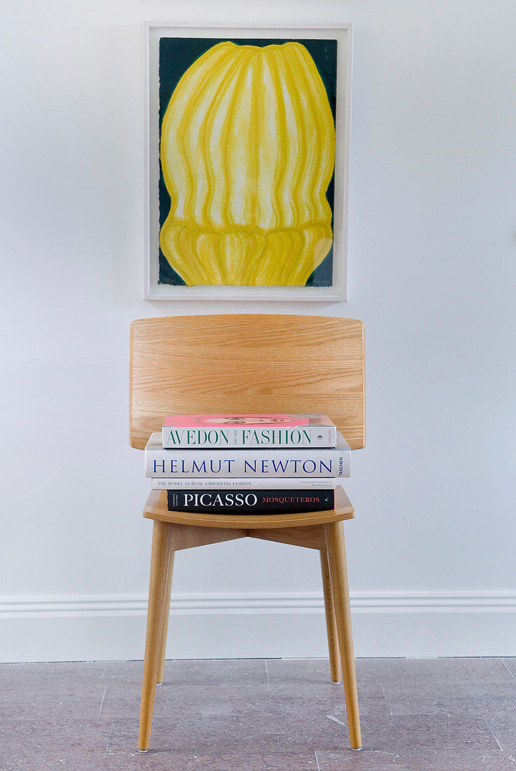 Books stacked on wooden chair below artwork on wall