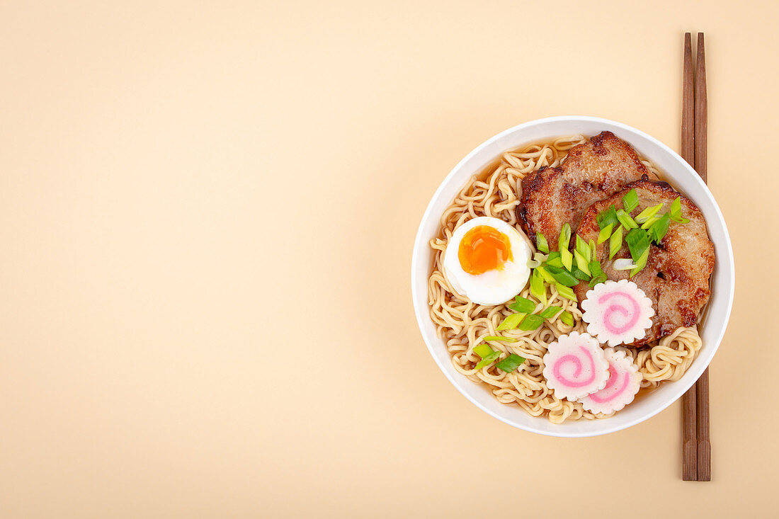 Top view of Japanese noodle soup ramen in white bowl with noodles, meat broth, sliced roasted pork, narutomaki, egg with yolk on pastel beige background