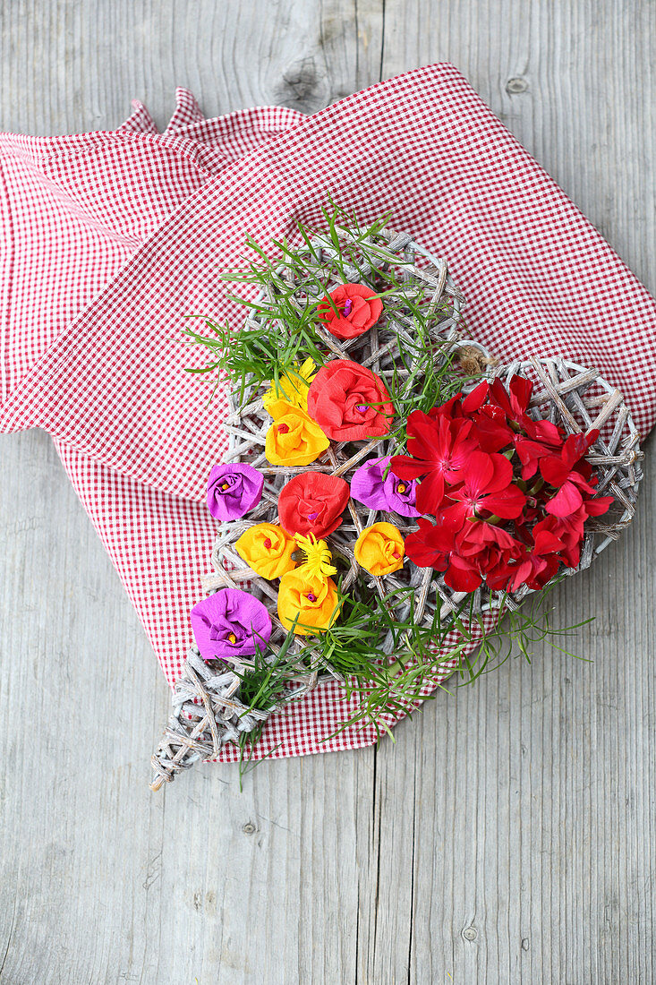 Wicker love-heart decorated with real geraniums and colourful paper flowers on gingham cloth