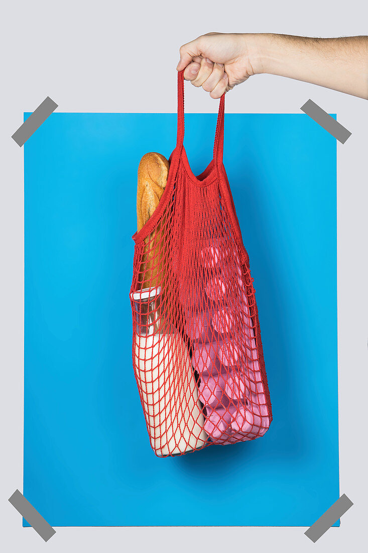 Hand carrying net bag with various groceries against blue rectangle during zero waste shopping