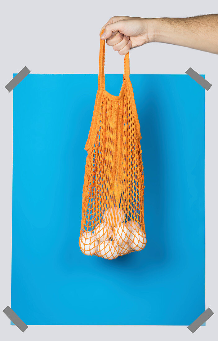 Hand carrying orange net sack with fresh eggs against blue rectangle during zero waste shopping