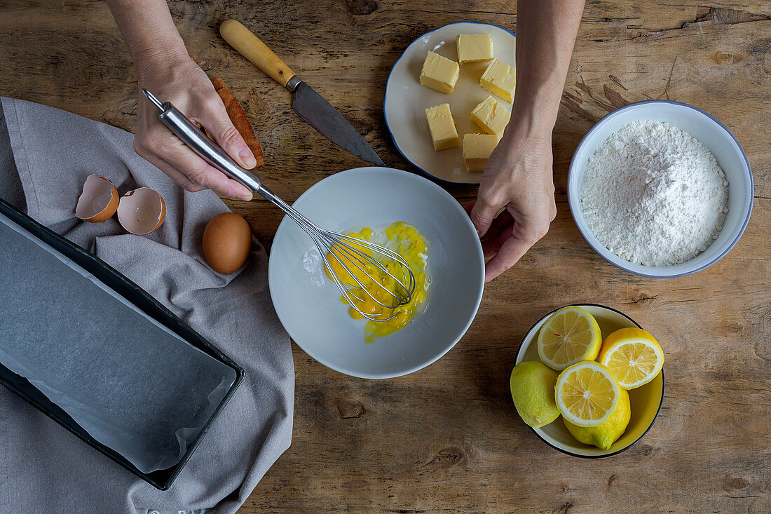 Whipping eggs in black bowl on wooden table with lemon, flour, butter and cinnamon sticks ingredients for cake