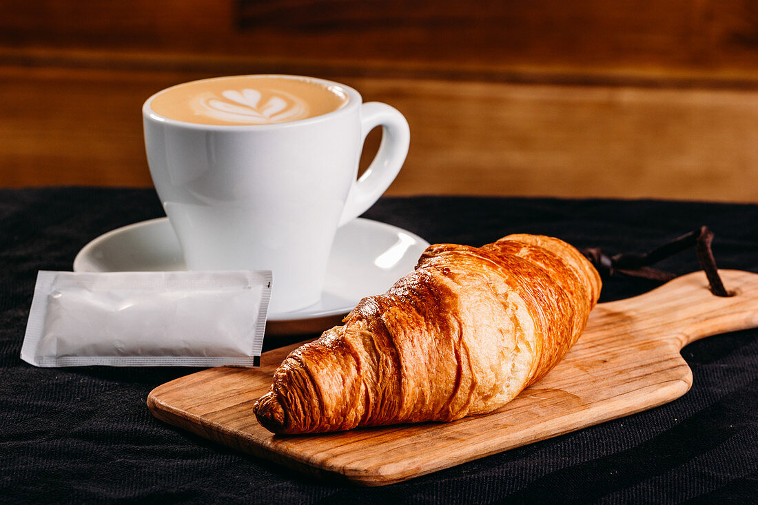 Golden crispy croissant served on wooden board with white cup of fresh coffee