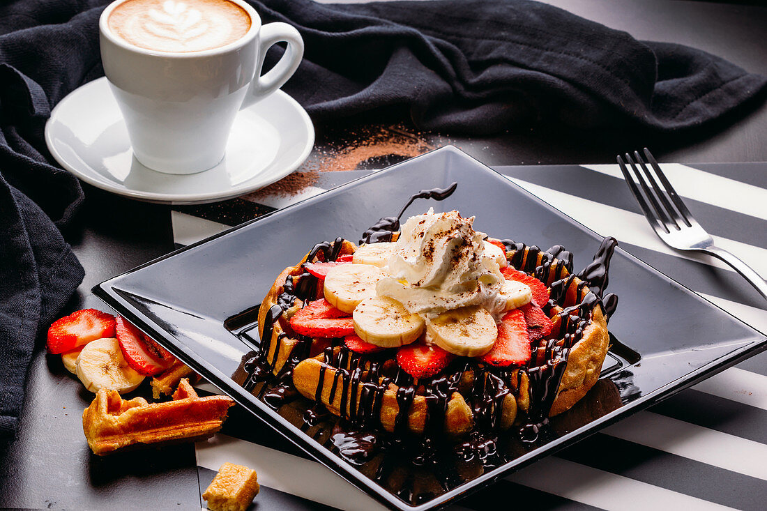 Cappuccino in white mug on table with plate of round waffle with banana and strawberry topped with chocolate sauce and whipped cream