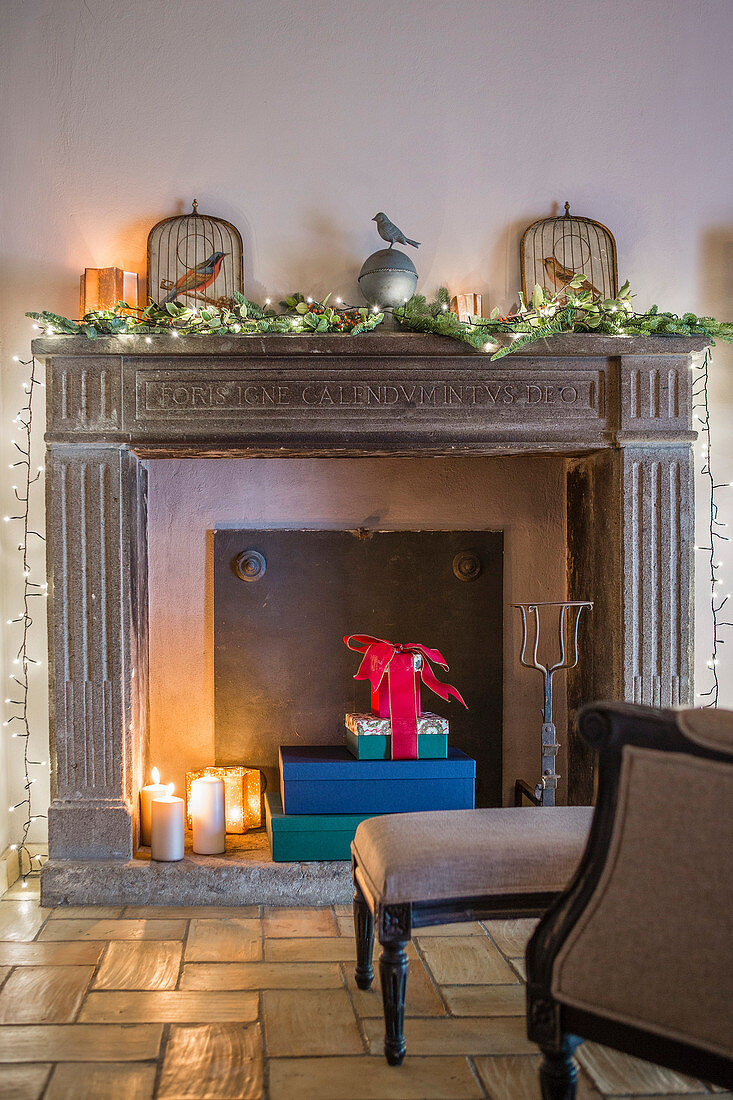Lit candles and wrapped gifts in disused fireplace with Christmas decorations on mantelpiece