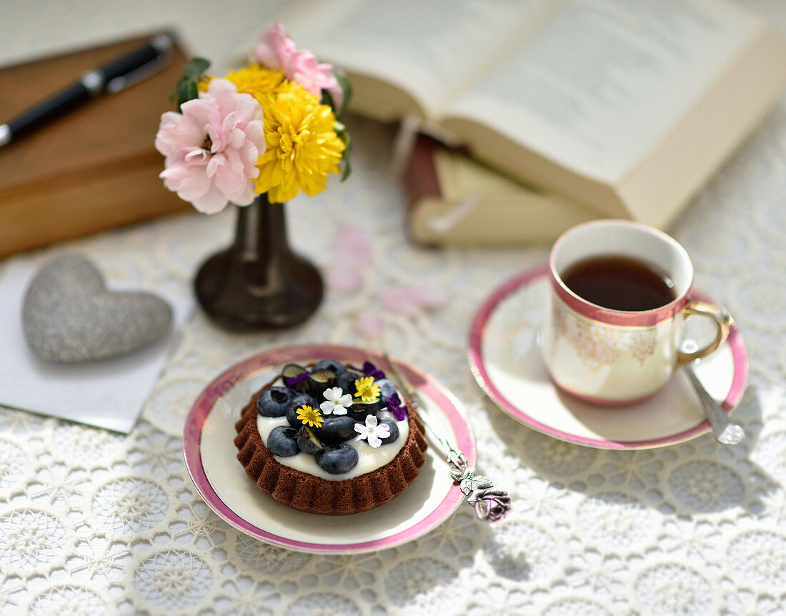 Veganes Heidelbeertörtchen zum Tee, im Hintergrund Blumen und Bücher