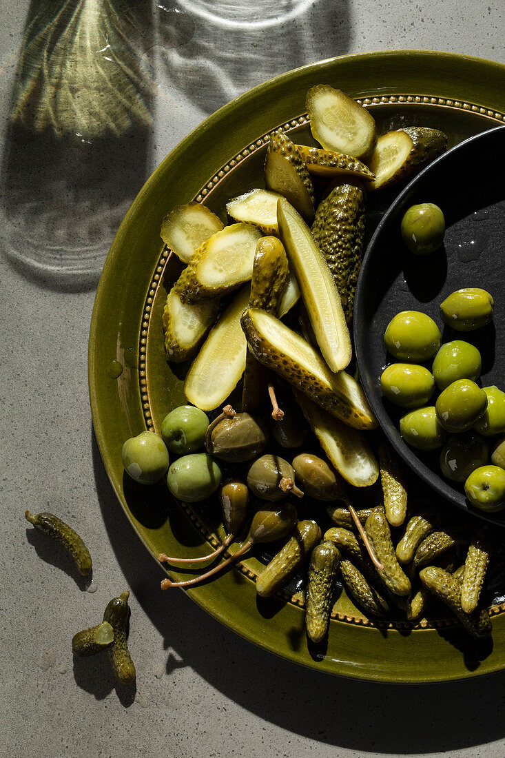 A selection of pickled gherkins, caper berries and green olives on an olive green plate