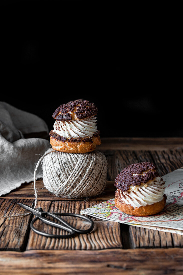 Homemade choux pastries with cream and chocolate scissors and ball of yarn arranged on texture wooden surface against black background