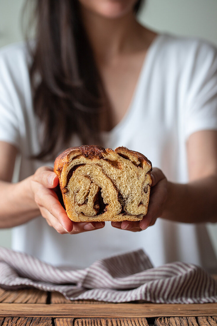 Crop female cook holding fresh twisted bread or cinnamon babka over wooden table with striped towel on blurred background