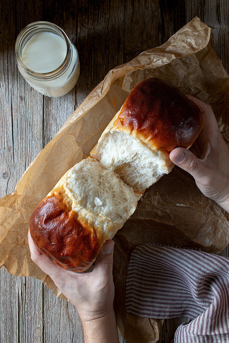 Top view of unrecognizable person dividing fresh bread buns over parchment near jar of milk on wooden table