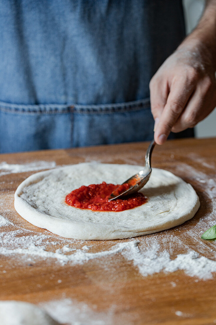 Anonymous chef smearing fresh tomato sauce on raw dough while cooking pizza on table