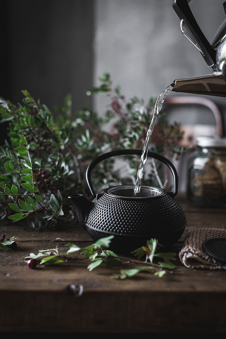 Round black teapot in hand of crop person pouring chamomile tea in black mug on wooden table decorating with green leaves