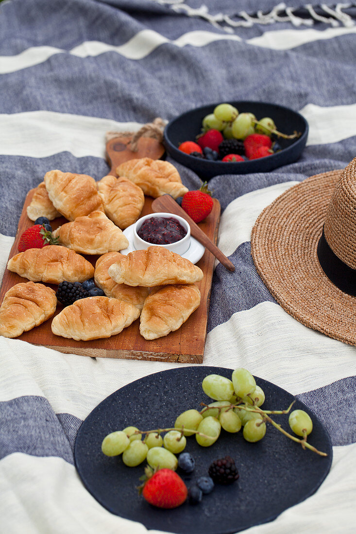 Picnic with berries, grapes, croissants and raspberry jam on a blue and white striped blanket