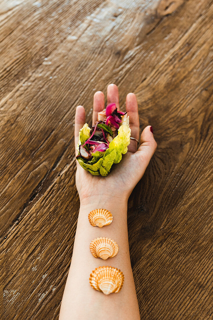 Hand of woman holding vegetarian rainbow lettuce wrap with seashells on wrist