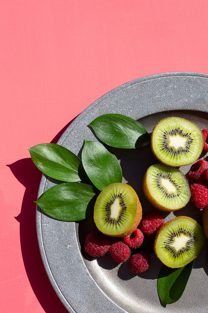 Kiwihälften mit Himbeeren auf Teller vor rosafarbenem Hintergrund