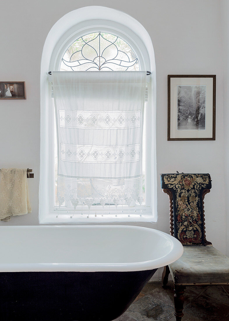 Embroidered, antique chair and lace curtain in arched window in vintage-style bathroom