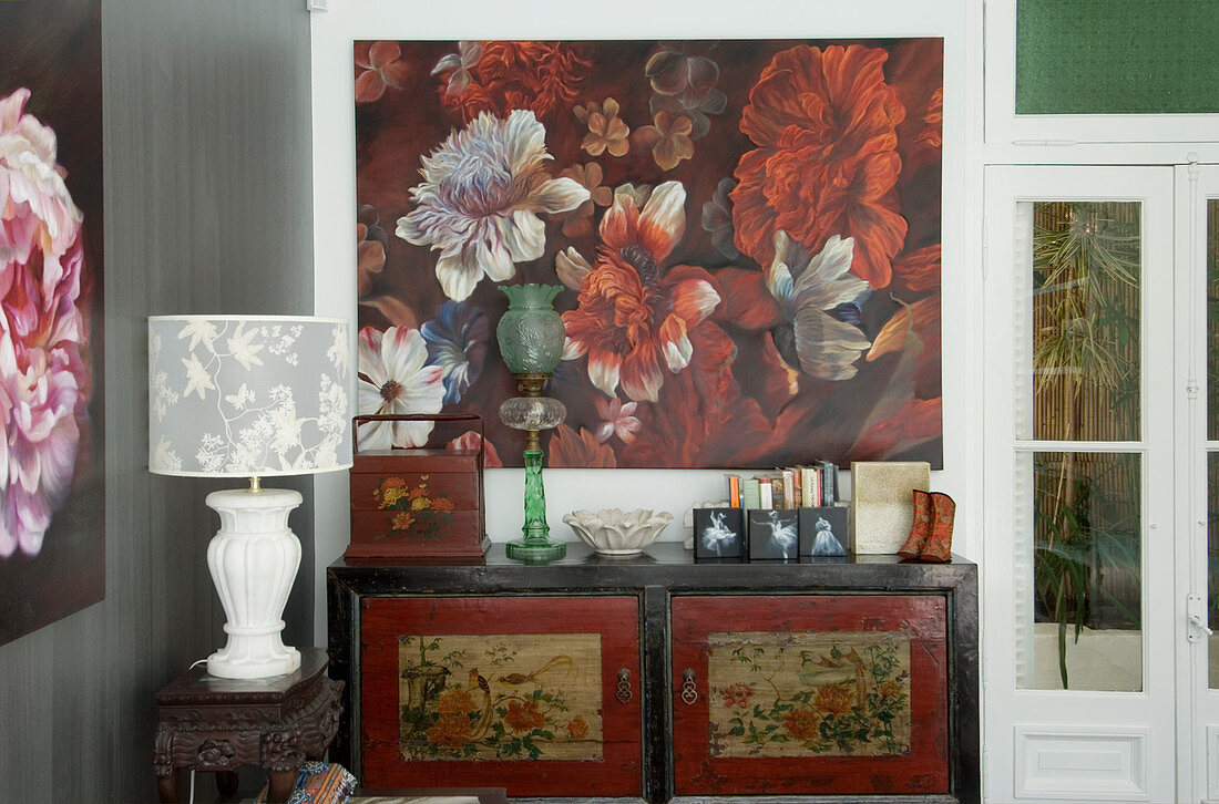 Oriental cabinet below floral artwork and table lamp on antique side table