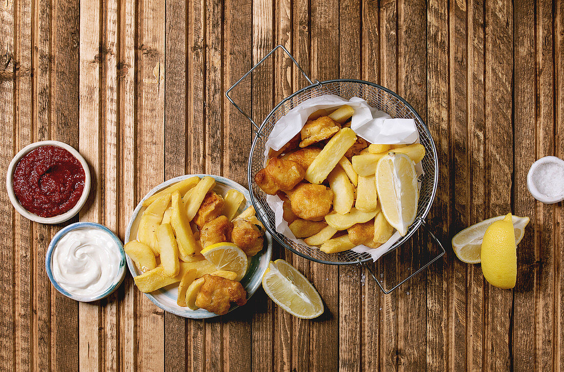 Classic british fast food fish and chips served in frying basket with lemons, red and white sauce, salt in ceramic bowls over wooden plank background